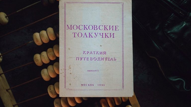 A vintage Moscow guidebook — "Moskovskie Tolkuchki" (Brief Guide, 1993) — rests on a worn abacus and dark leather, photographed from above in soft, moody light. The faded paper, purple typography and subtle stains tell a quiet story of markets, travel and everyday life, while the textured beads and cracked cover evoke tactile nostalgia and the charm of analog objects.