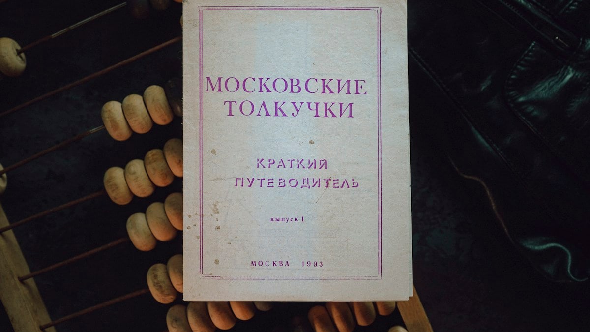 A vintage Moscow guidebook — "Moskovskie Tolkuchki" (Brief Guide, 1993) — rests on a worn abacus and dark leather, photographed from above in soft, moody light. The faded paper, purple typography and subtle stains tell a quiet story of markets, travel and everyday life, while the textured beads and cracked cover evoke tactile nostalgia and the charm of analog objects.